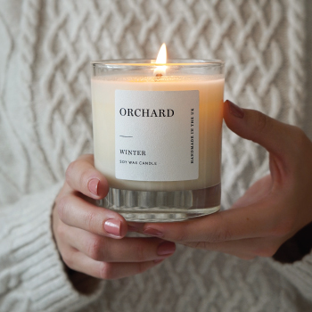 Woman holding lit candle. An image showing square textured white labels on an Orchard Candle products.